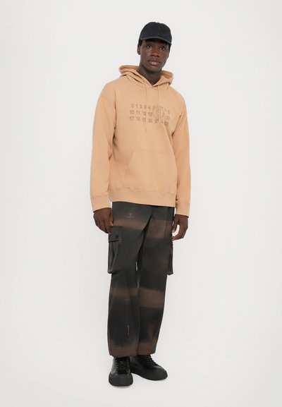 Beige hoodie med framficka, grafiska detaljer i brunt. Kombinerat med svarta cargobyxor med flera sidofickor och ett gradientmönster. Svarta skor.