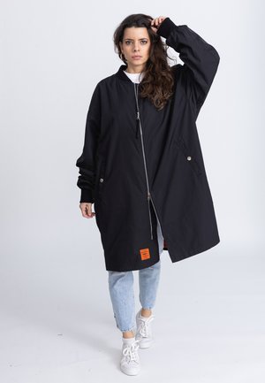 Zwarte lange jas met ritssluiting aan de voorkant, verlaagde schouders en zijzakken. Kenmerkt zich door een oranje label op de zoom en is gecombineerd met lichte jeans en witte sneakers.