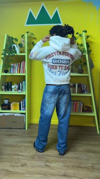 Λευκό φούτερ με κουκούλα με την επιγραφή «FOREVER FORWARD 2025 BORN TO RACE» σε κόκκινο, σε συνδυασμό με ξεβαμμένο μπλε τζιν. Το φόντο περιλαμβάνει πράσινα ράφια και κίτρινο τοίχο.