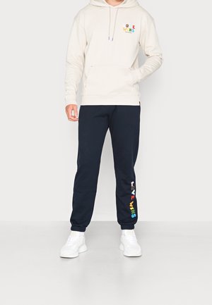Felpa crema con tasca frontale, con la scritta "LOVE WINS" in lettere colorate. Abbinata a pantaloni della tuta blu navy che mostrano la stessa scritta in colori dell'arcobaleno.