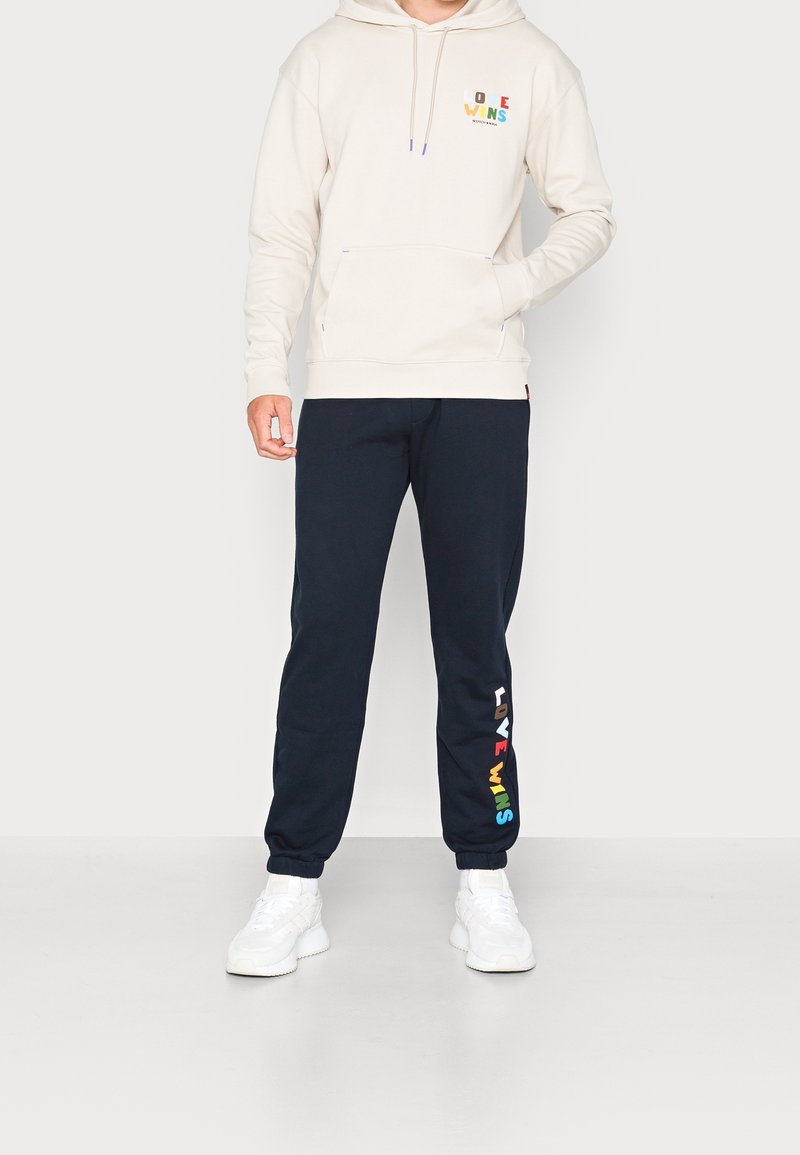 Felpa crema con tasca frontale, con la scritta "LOVE WINS" in lettere colorate. Abbinata a pantaloni della tuta blu navy che mostrano la stessa scritta in colori dell'arcobaleno.