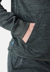 Il pullover grigio con texture presenta una tasca a marsupio, polsini a costine e una piccola bandiera svedese come dettaglio sull'orlo. Il materiale sembra morbido e flessibile.