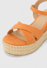 Oransje semsket skinn sandaler med kryssende stropper og en vevd jute plattform såle. Glatt overflate med rund tå og flat hel.