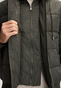 Giacca bomber verde imbottita con zip frontale, due tasche laterali e cappuccio. Presenta una texture trapuntata e polsini elastici.
