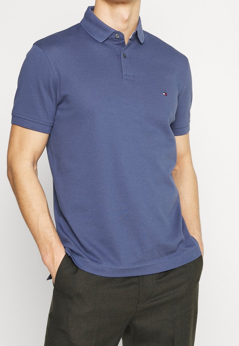 Man med en blå kortärmad pikétröja med en liten logotyp på bröstet och mörka byxor, händerna nedstuckna i fickorna.