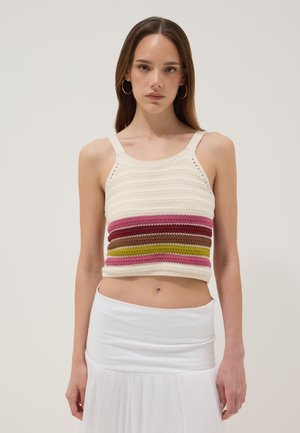 Jonge vrouw met lang bruin haar, die gouden oorringen draagt, een mouwloze gestreepte gehaakte cropped top en een witte rok, staand tegen een effen achtergrond.