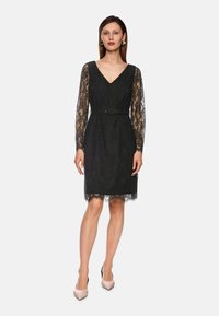 Schwarzes Spitzenkleid mit langen Ärmeln, V-Ausschnitt und tailliertem Bund. Verfügt über einen wellenförmigen Saum und ein zartes Spitzenmuster über das gesamte Kleid.