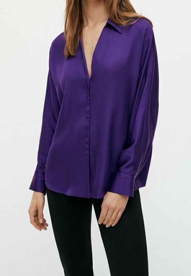 Femme portant un chemisier violet à manches longues, boutonné avec un col, et un pantalon noir, sur un fond clair uni.