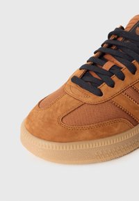 Sapatilha em camurça castanha e malha, com atacadores pretos, parte superior texturizada e sola de borracha com borda serrilhada e padrão subtil.