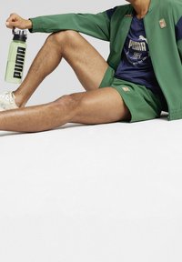 Grön träningsjacka och shorts, blå skjorta med logotyp, krämfärgade sneakers. Håller en genomskinlig grön vattenflaska med svart branding.