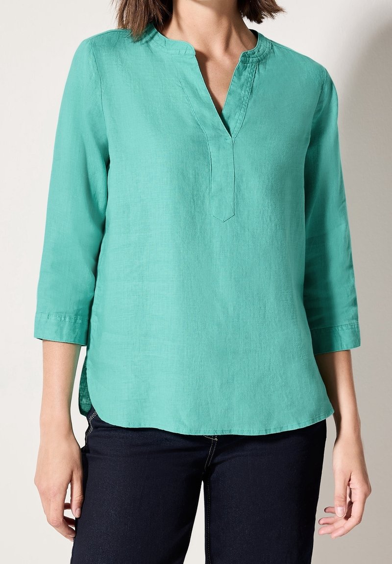 Femme portant un chemisier turquoise à manches longues avec une encolure fendue et un pantalon foncé, montrée des épaules aux hanches.