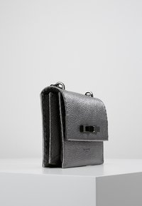 Szara teksturowana torebka na klapie, z czarnym zapięciem i srebrnymi elementami, stoi pionowo na białej powierzchni.