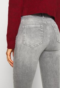 Jeans de mezclilla gris ajustados con un sutil desgastado, bolsillos traseros y una cintura alta, combinados con un suéter de punto rojo.