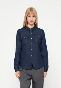 Camisa de ganga escura de manga longa com fecho dianteiro de botões, colarinho pontudo e dois bolsos no peito. Textura suave com um design ligeiramente ajustado.