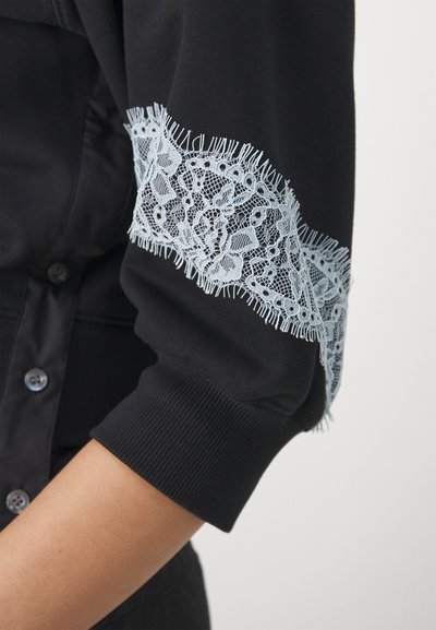 Maglione nero con bordi in pizzo azzurro chiaro sulle maniche, rifinito con frange. Il tessuto ha una texture liscia e polsini a coste.