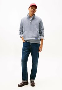 Pullover in pile grigio con collo a mezza zip e tasca frontale, abbinato a jeans in denim blu scuro e mocassini marrone scuro. Il berretto rosso aggiunge colore.