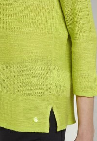 Pull vert citron en maille à large tissage, manches trois-quarts et fente latérale. Il présente un bouton rond et brillant sur l'ourlet.