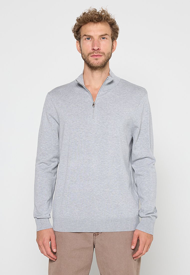 Man met krullend haar, gekleed in een lichtgrijze sweater met kwart rits en beige broek, staand met een neutrale uitdrukking tegen een witte achtergrond.