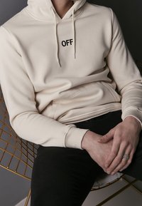 Osoba v krémovém mikině s textem "OFF", černých kalhotách, sedící na zlaté drátěné židli s rukama sepjatýma, nosící stříbrný prsten.