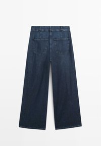 Donkerblauwe wijd uitlopende spijkerbroek met twee achterzakken en riemlussen, van achteren weergegeven op een witte achtergrond.