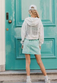Grijze hoodie met tekst op de rug, gecombineerd met een mintgroene spijkerrok, witte sokken en grijze sneakers, tegen een teal deur.