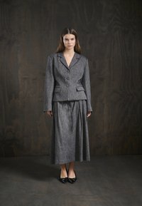 Grauer taillierter Blazer mit zwei Knöpfen, kombiniert mit einem langen, passenden Rock. Der Stoff hat eine dezente Textur. Schwarze spitze Absatzschuhe runden den Look ab.