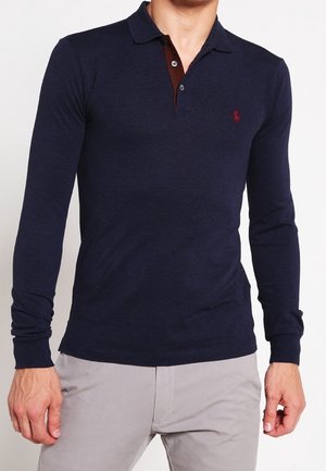 Man draagt een nauwsluitend donkerblauw poloshirt met lange mouwen, een bruine knopenstrook en lichtgrijze broek, staand met ontspannen armen.