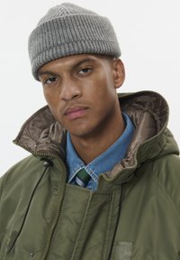 Zaļa neilona parkas ar bežīgu oderi, uzvilkta pār zilu kreklu un kaklasaiti. Pelēka ribota cepure. Tuvplāns uz sejas iezīmēm, neitrāla izteiksme.