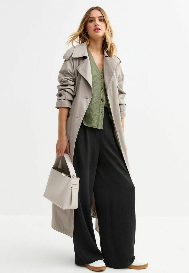 Licht beige trenchcoat over een groene button-down shirt, gecombineerd met zwarte wijde broek en witte sneakers. Houdt een lichtgrijze handtas vast.
