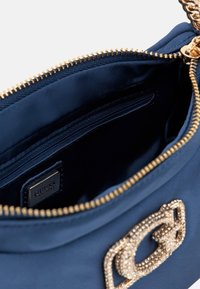 Interno di una borsa a tracolla in tessuto blu navy con zip dorata, dotata di un'etichetta con logo e di una tasca con zip per piccoli oggetti. Presenti dettagli metallici.