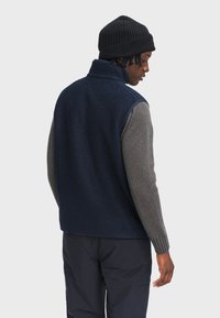 Gilet in fleece blu navy con maniche in maglia grigia, colletto alto e polsini a coste. Indossato sopra pantaloni scuri, visto da dietro.