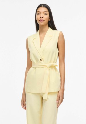 Donna con capelli lunghi e scuri che indossa un blazer senza maniche giallo pallido con cintura e pantaloni larghi abbinati, in piedi davanti a uno sfondo bianco.