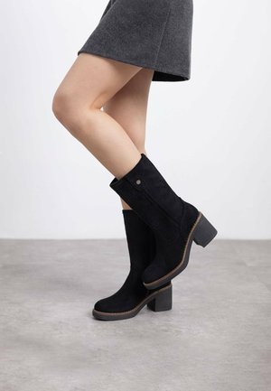 Botas de tobillo de ante negro con acabado texturizado, punta redondeada y tacón grueso. Presenta cremallera lateral y un detalle de costura clara en contraste.