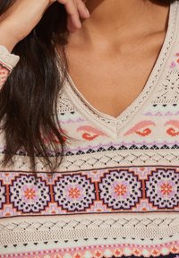 Pull en tricot avec un col en V, tissu beige orné de motifs multicolores, comprenant des cercles et des vagues, avec des trous décoratifs le long de l'encolure.