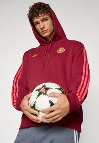 Maroon hoodie with a front pocket, featuring a gold emblem and pink-striped sleeves. Model holds a white and black star-patterned soccer ball.