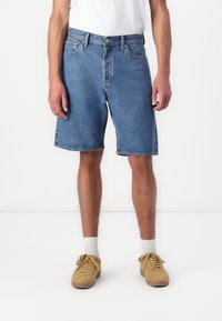 Shorts en denim bleu clair avec une coupe droite, un design à cinq poches et un ourlet brut. Porté avec des baskets beiges et des chaussettes blanches.