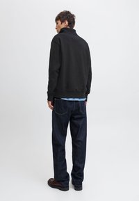 Mees seisab seljaga, kandes musta pulloverit, tumelisi teksapükse, pruune kingi ja helesinist särk silma torgib pulloveri alt.