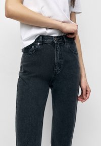 Persona que lleva jeans de tiro alto negros y una camiseta blanca, ajustando la cintura con una mano contra un fondo liso.