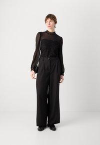 Zwarte kantblouse met doorzichtige puffmouwen, hoge halslijn en brede zwarte broek. Een slanke zwarte handtas rondt de outfit af.