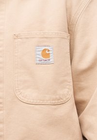 Beige arbejdsjakke med lomme og hvid Carhartt-mærkat med orange logo og rød tekst.
