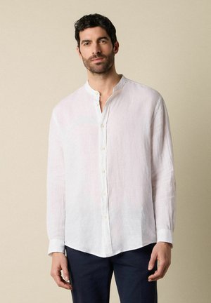 Man draagt een wit linnen overhemd met lange mouwen en mandarijnkraag, gecombineerd met een donkere broek, staand tegen een beige achtergrond.