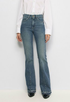 Mujer que lleva vaqueros azules de talle alto con aberturas delanteras en el dobladillo, combinados con una camisa blanca abotonada y zapatos negros puntiagudos.