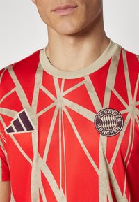 Jersey rojo de manga corta con un patrón geométrico beige, cuello redondo, un logo de Adidas en azul oscuro y un escudo bordado del FC Bayern München.