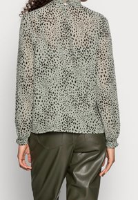 Blusa verde transparente con patrón de puntos negros, puños fruncidos y cierre trasero con botones, combinada con pantalones de cuero verde oliva.