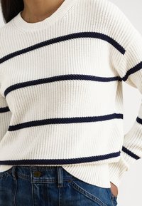 Pletený sveter v krémovej farbe s námorníckymi horizontálnymi pruhmi. Má okrúhly výstrih a rebrovanú textúru, doplnený o rifle v páse.