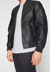 Schwarze Lederbomberjacke mit strukturiertem Finish, ausgestattet mit einem Reißverschluss vorne, seitlichen Reißverschlusstaschen, gerippten Bündchen und einem schlichten grauen T-Shirt darunter.