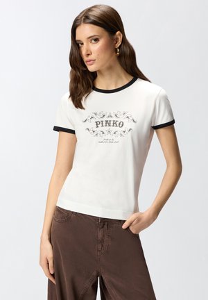 Mujer con camiseta blanca con ribete negro y texto "PINKO", combinada con falda marrón de talle alto, posando con una mano en el bolsillo.