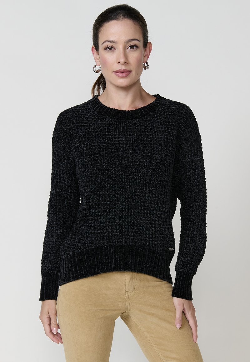 Črn strukturiran pulover s okroglo kakčko, z ribastimi manšetami in robom. Nosijo ga z svetlo rjavimi kordnimi hlačami, ki imajo sproščen kroj.