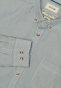 Koszula z długimi rękawami, zapinana na guziki, w jasno-niebieskie i białe paski. Posiada brązowe guziki, kieszeń na piersi oraz mankiety złożone w stylu.