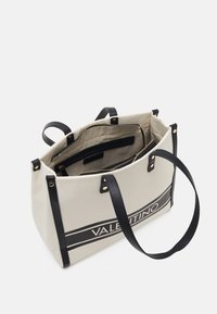 Canvasväska i beige med svarta läderremmar och detaljer. Har ett blixtlåsfack och tryckt varumärke på framsidan.